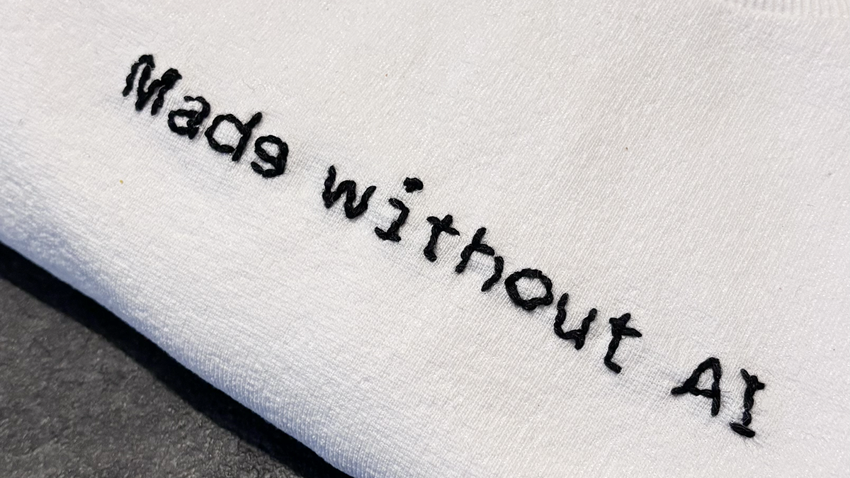Scritta ‘Made without AI’ ricamata a mano in nero su tessuto bianco, con lettere imperfette e irregolari a evidenziare l’esecuzione manuale.